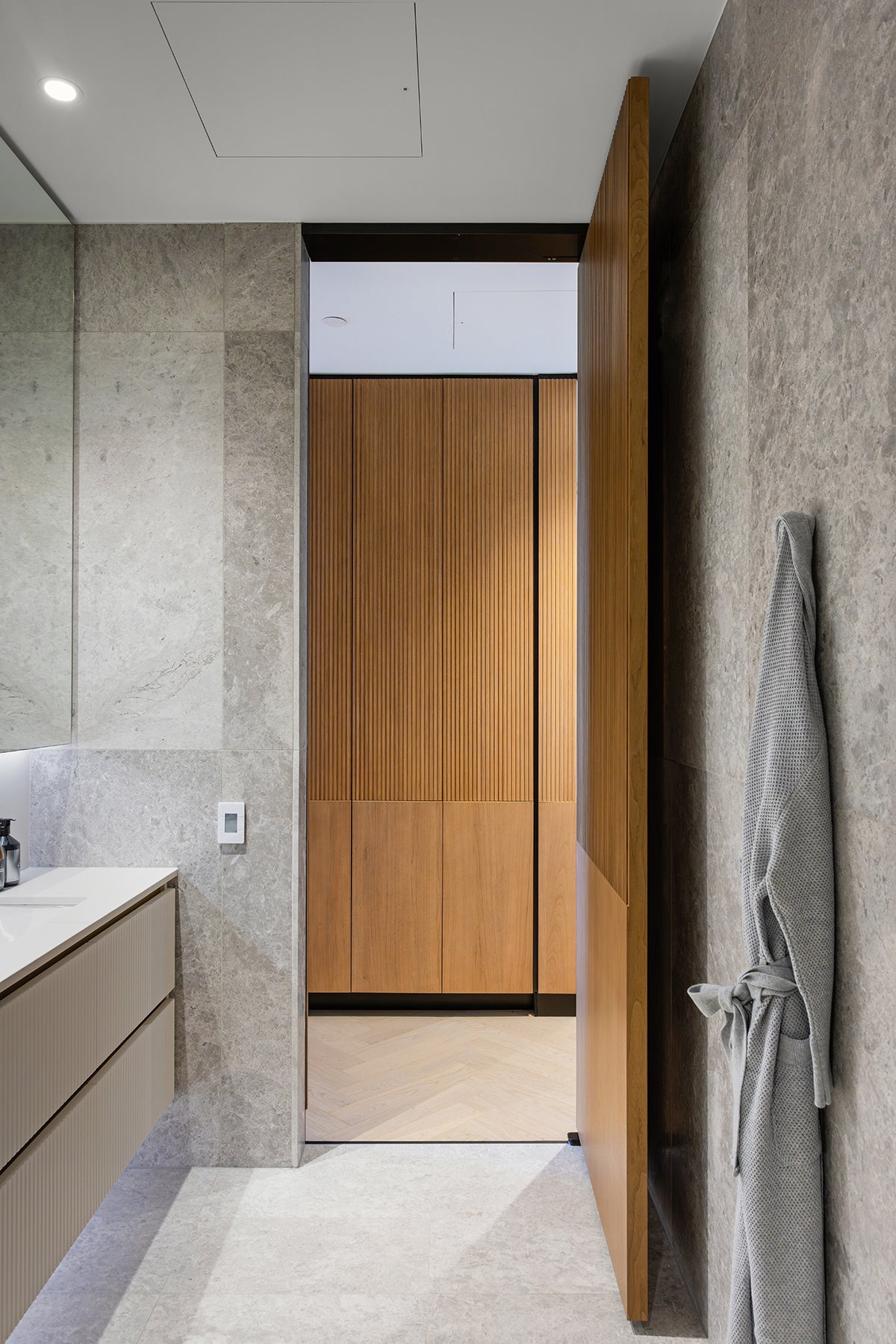 A modern bathroom interior with a gray stone-textured wall, a gray robe hanging on the right, a light wood vanity on the left, and an open wooden door revealing a wood-paneled closet in the background.