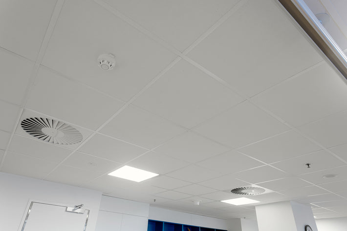 White ceiling tile grid with recessed square lights, circular ventilation grilles, and a smoke detector in an indoor office or commercial space.