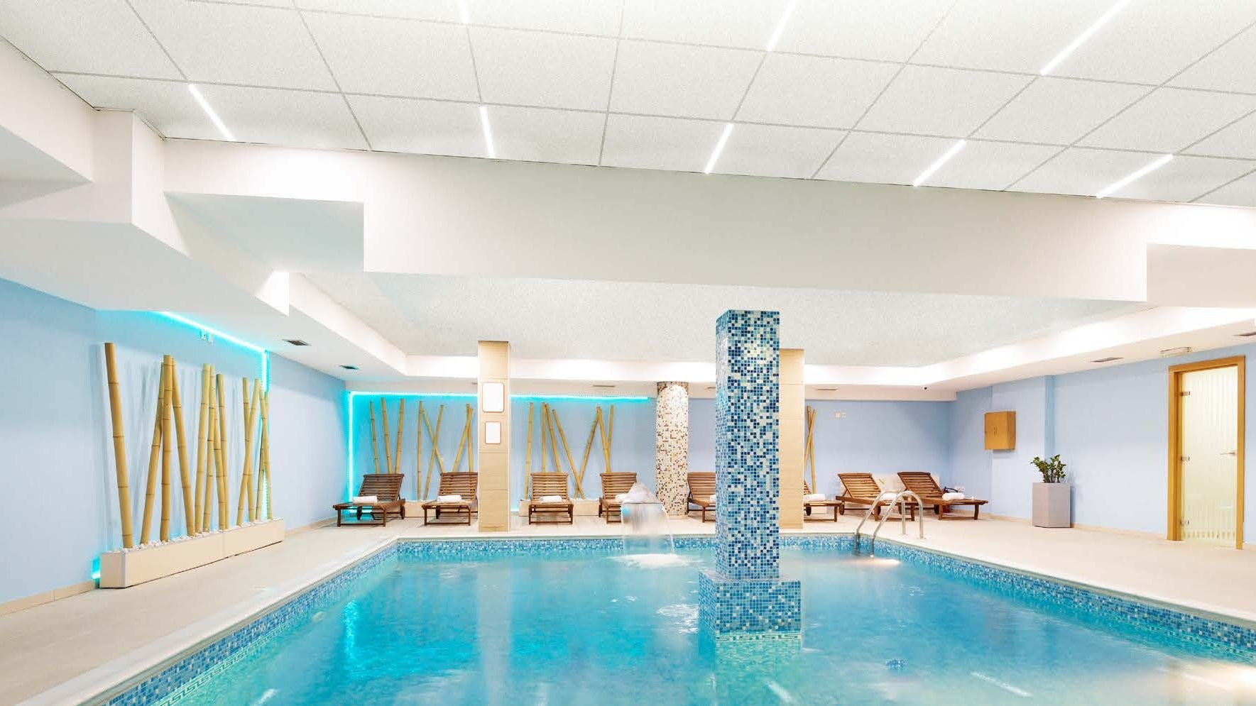 An indoor swimming pool with light blue walls, a central mosaic blue column, lounge chairs, and a modern ceiling with integrated kwiklux lighting.