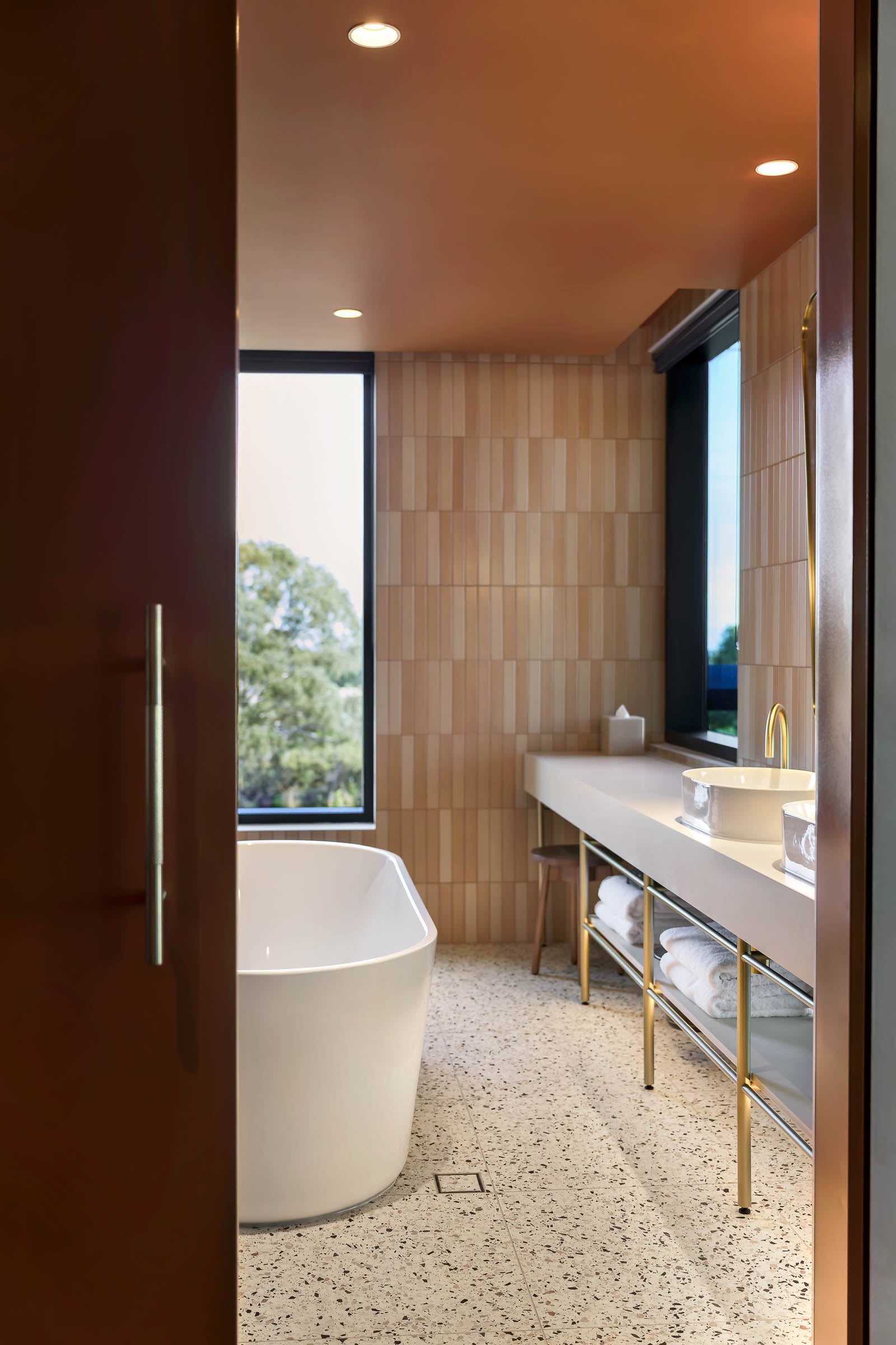A modern bathroom interior with a freestanding white bathtub, a long white vanity with gold metal legs, terrazzo flooring, and light wood wall panels. A dark wood door frame is visible on the left, and windows look out to trees.