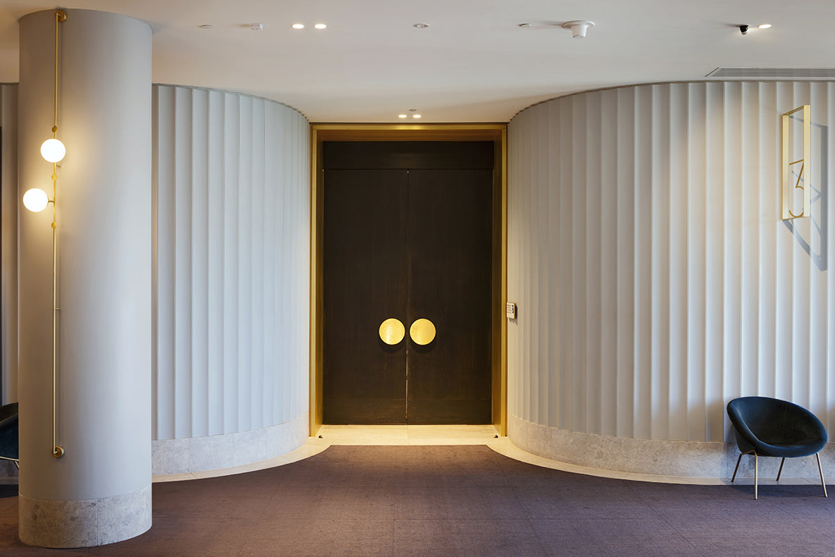 A modern interior hallway with curved white fluted walls, a dark double door with two round gold handles, a tall cylindrical wall lamp on the left, and a black chair on the right.