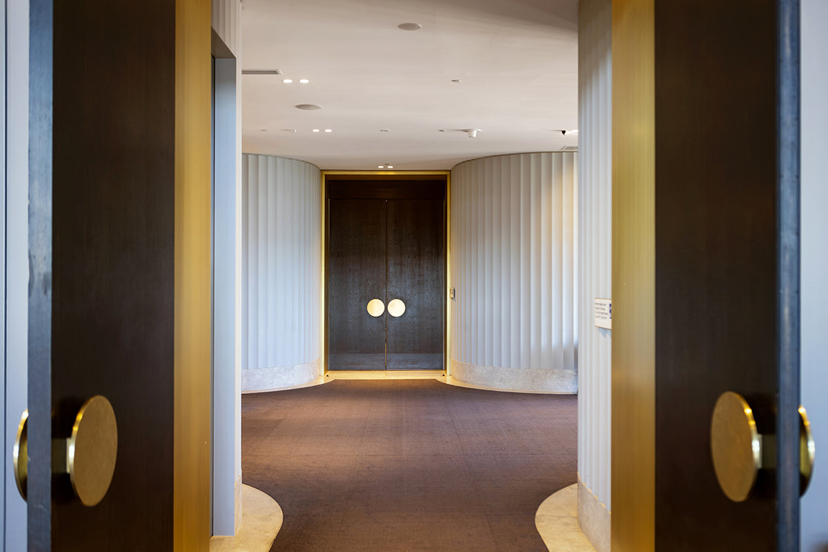 A view through open dark wood doors into a modern interior hallway with curved white ribbed walls, a dark wood double door at the end, and gold round door handles.
