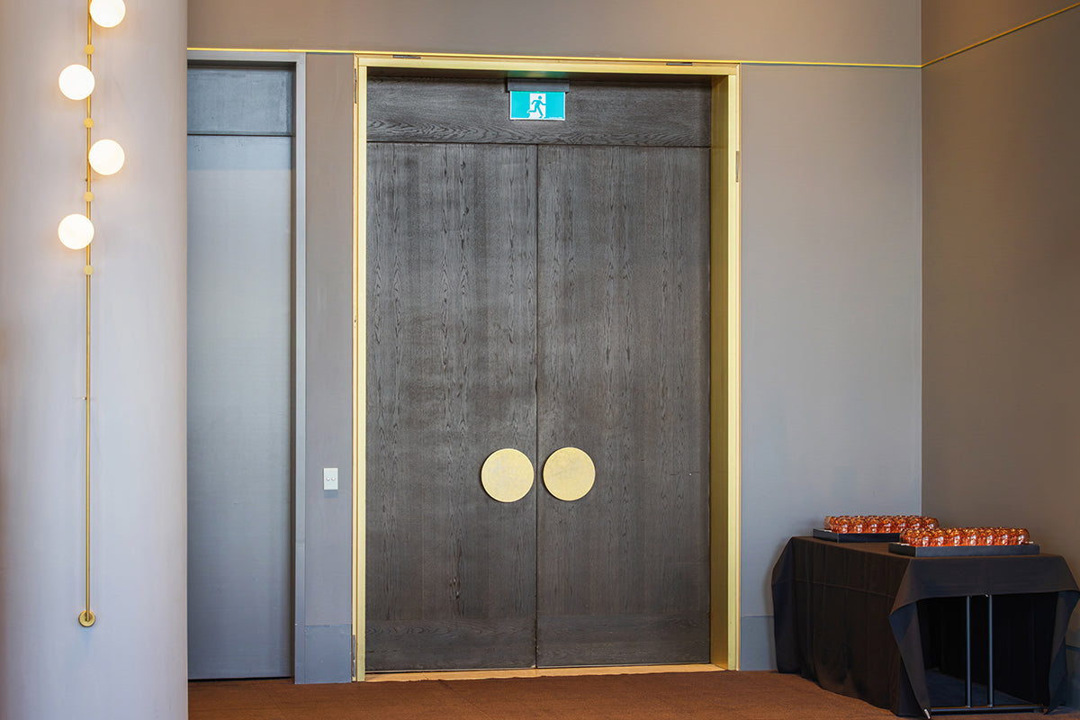 A pair of dark gray textured double doors with circular gold handles, set within a gold-trimmed frame in a modern interior hallway.